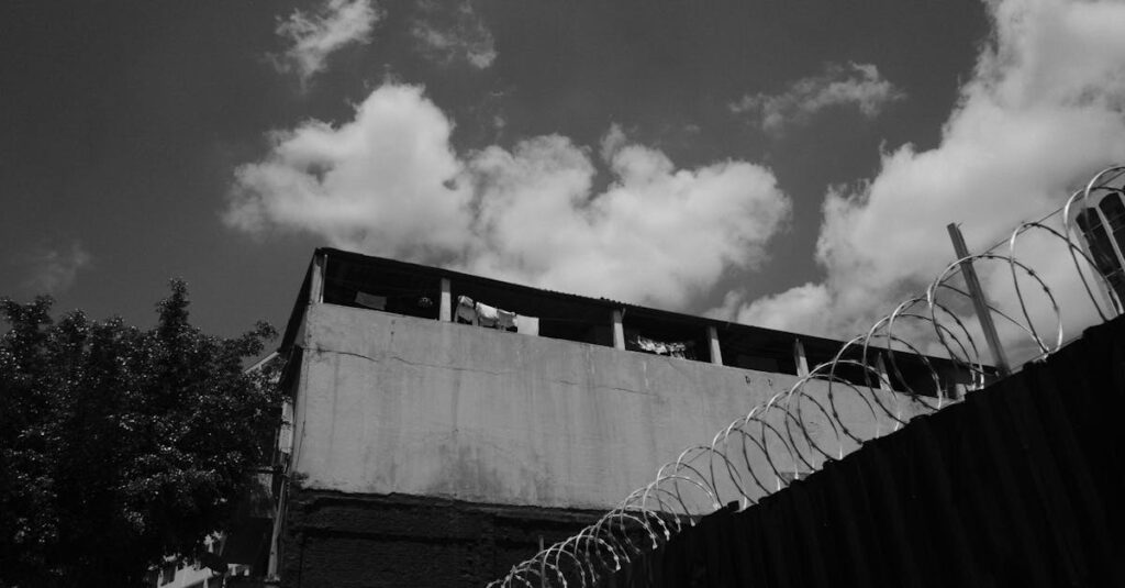 Black and white photograph of an urban building with barbed wire fence under a cloudy sky.