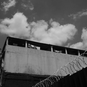 Black and white photograph of an urban building with barbed wire fence under a cloudy sky.